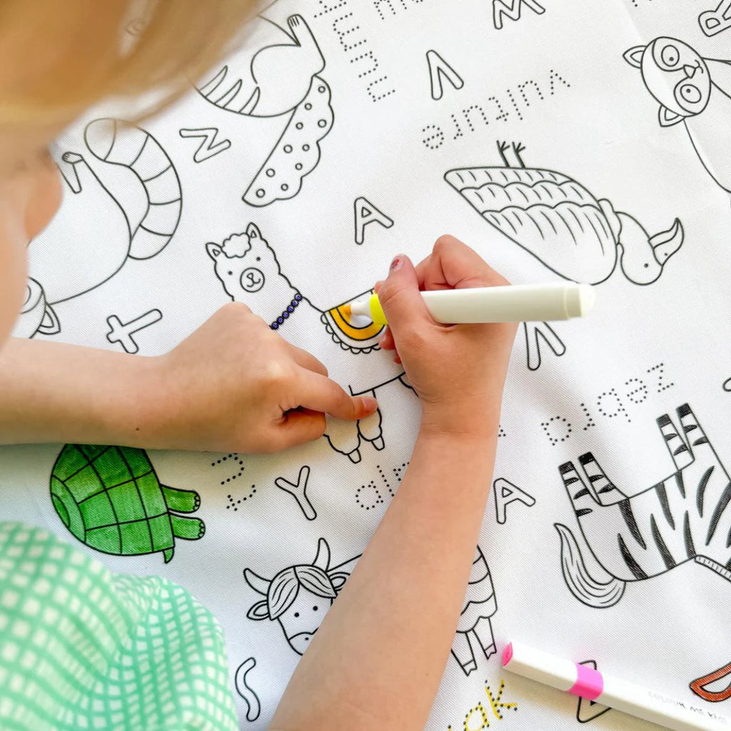 Close-up of child’s hands using washable markers to colour Alphabet Animals tablecloth, featuring outlined animal letters like zebra, giraffe, and elephant.