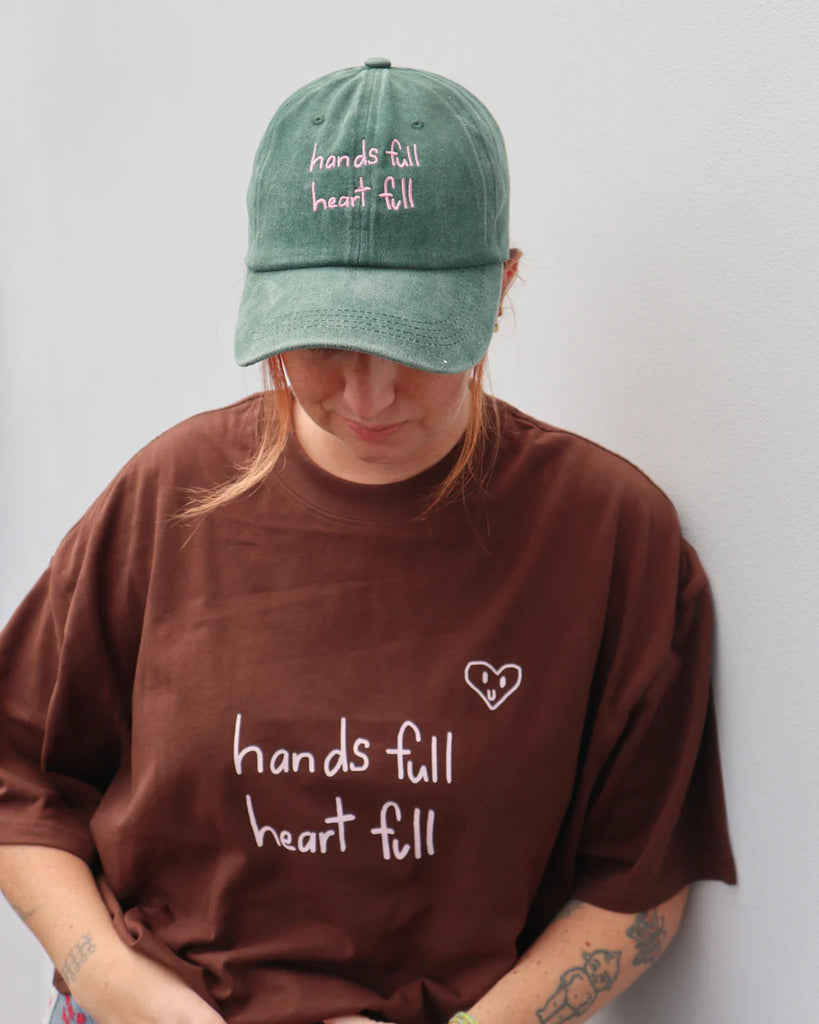 Person wearing brown shirt and green "Hands Full Heart Full" cap.