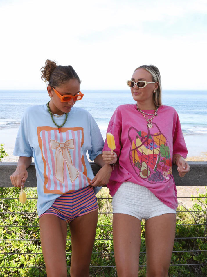 Two women wearing matching oversized t-shirts, one hot pink with fruit market graphic and the other light blue with bow graphic, standing side by side outdoors