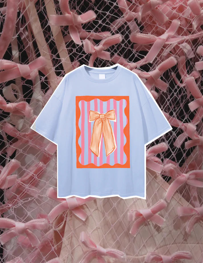 Light blue oversized t-shirt with orange and pink bow print, laid flat on a pink mesh background