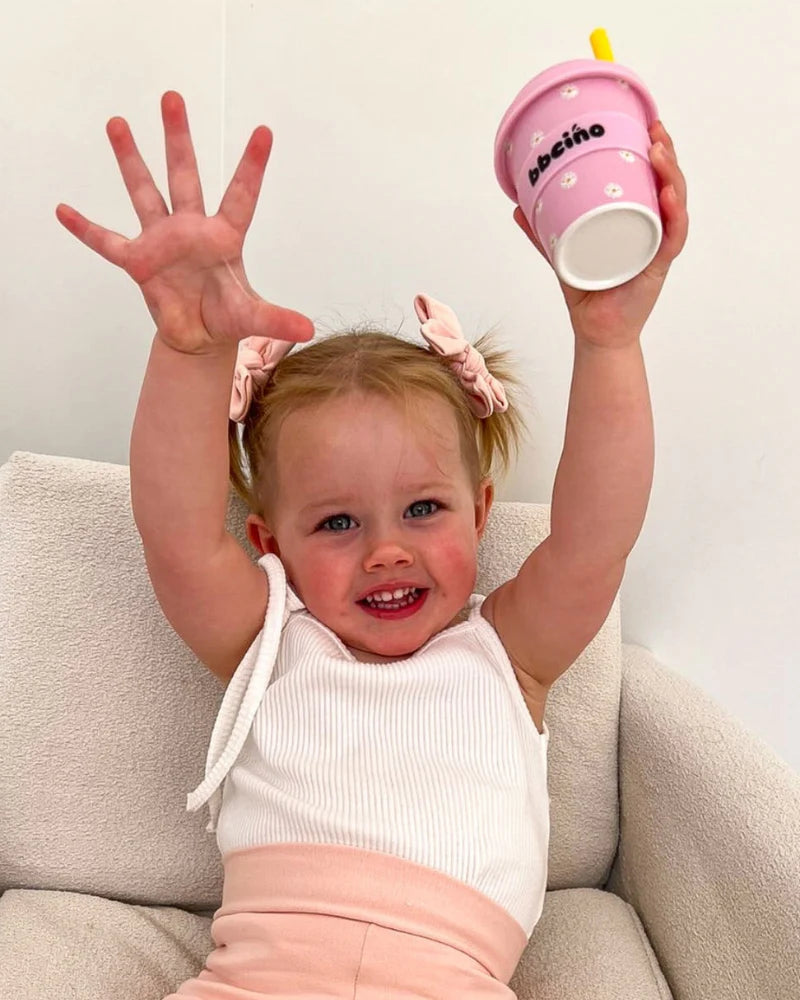 Smiling toddler holding a BBCino pink daisy cup with yellow straw, celebrating eco-friendly fun.