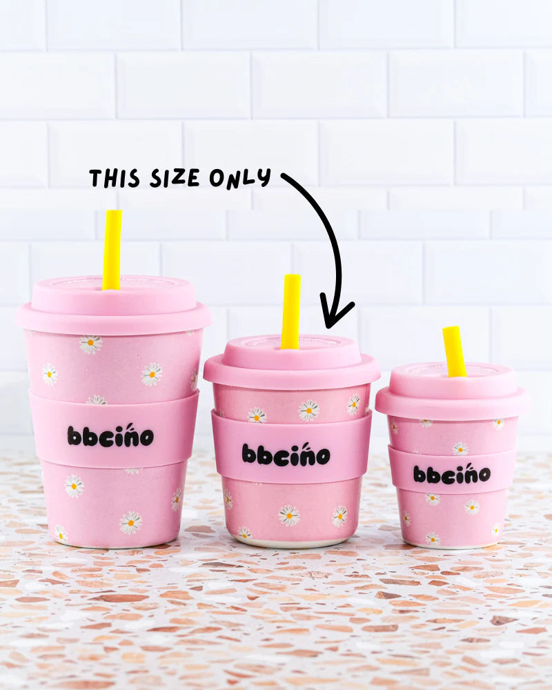 Three BambinoCino pink reusable cups with daisy design and yellow straws lined up against a tiled wall.
