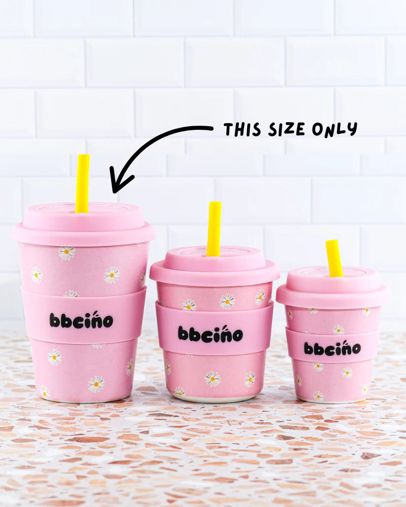 Three BiggieCino pink reusable cups with yellow straws lined up against a white tiled wall.