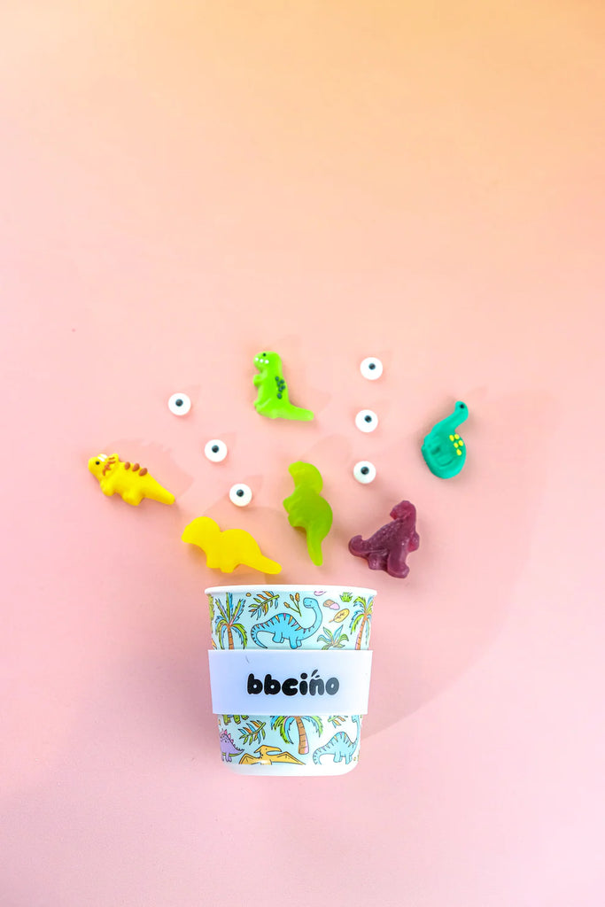 BBCino reusable cup 240ml in Sipasaurus dinosaur design, styled with toy and lolly dinosaurs and sprinkles on pink background, sustainable kids’ keep cup.