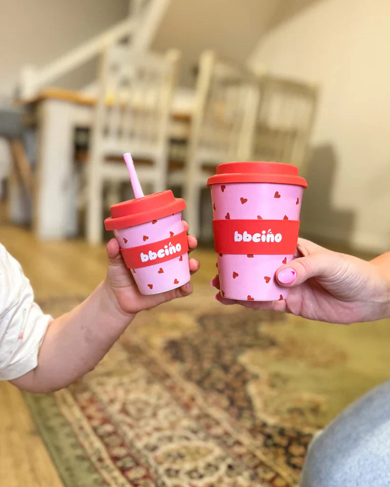 2 people "cheers"ing reusable cups with pink and red love heart design.