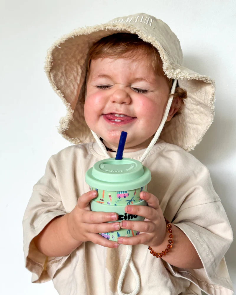 Child pictured with Bbcino 240ml reusable cup with beach design on mint green background,with mint green lid and navy coloured straw. 