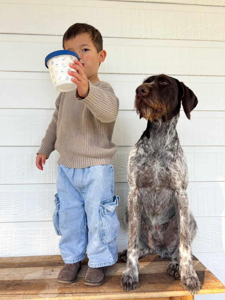 Child standing with dog holding BBCino reusable bamboo snack pot in Brunch-Kins print with navy silicone lid, eco-friendly snack container for families.