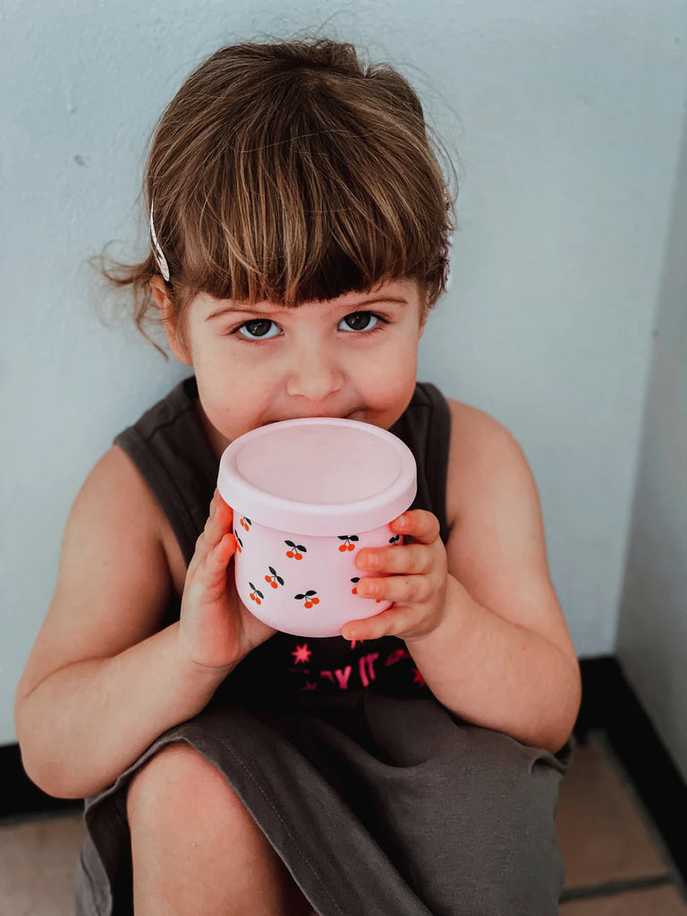 Child holding BBCino reusable silicone snack pot in Cherub pink cherry design, 250ml unbreakable and leak-resistant food container for kids’ snacks.