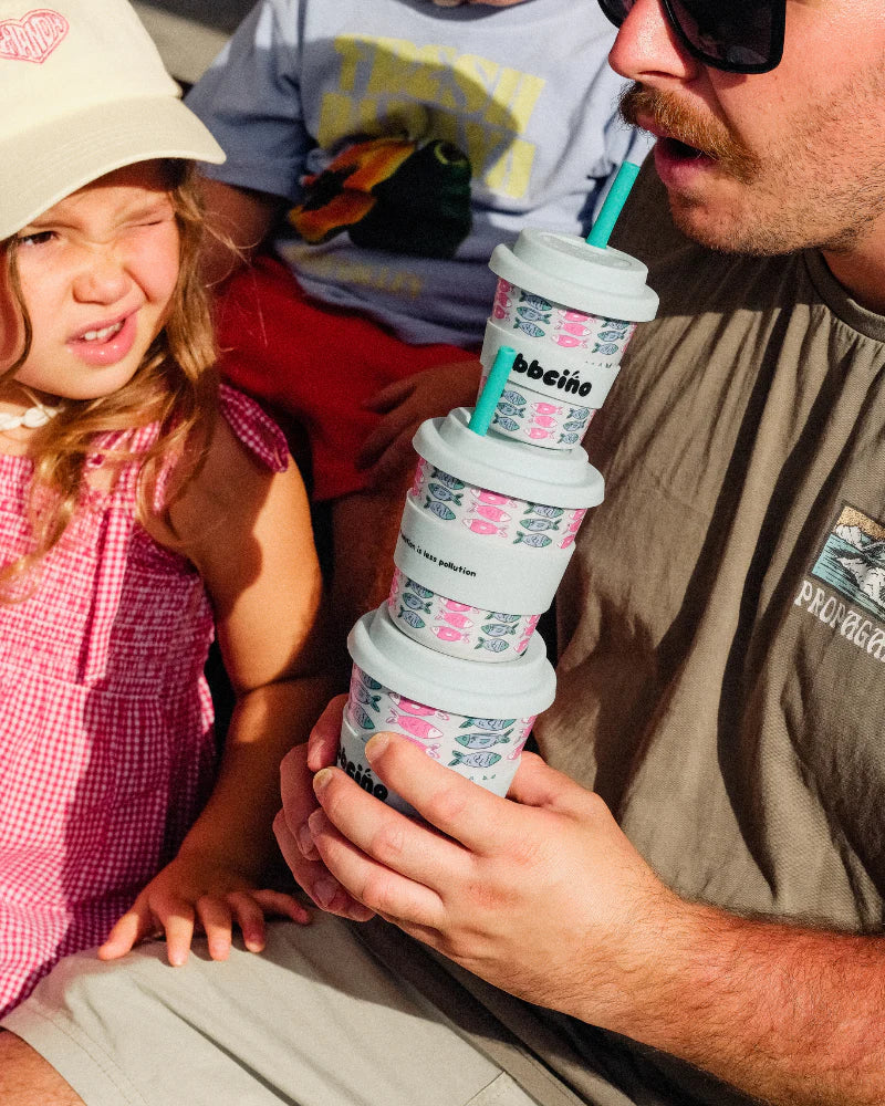 Adult holding stack of fish-patterned BBCino BiggieCino cups while smiling child looks on
