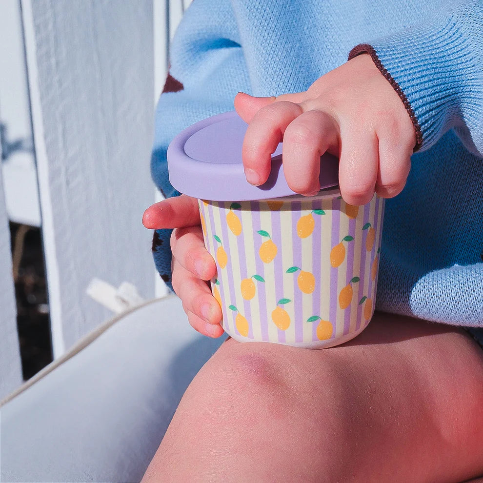 Child holding BBCino bamboo reusable snack pot with lemon print and purple silicone lid, dishwasher-safe eco-friendly container for kids’ snacks.