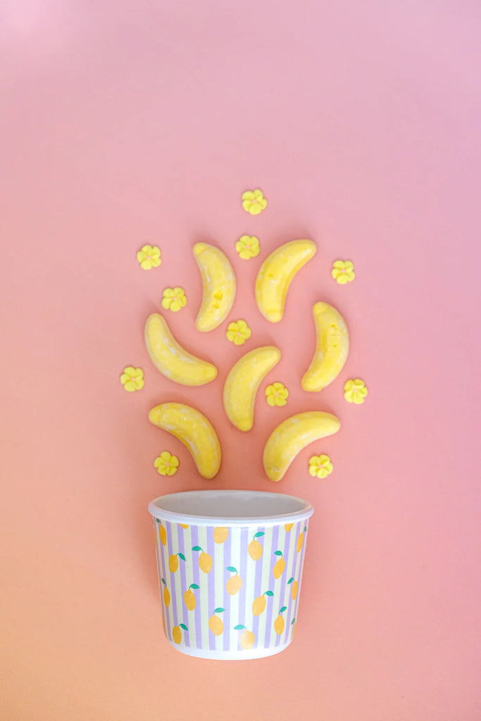 BBCino bamboo reusable snack pot with lemon design and lilac lid, styled with lolly bananas and popcorn on pink background, sustainable kids lunchbox container.