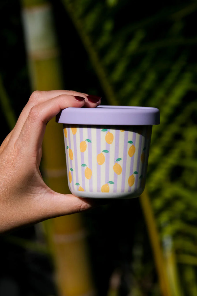 Hand holding BBCino reusable bamboo snack pot with lemon print and lilac silicone lid, sustainable eco-friendly container for snacks on the go.