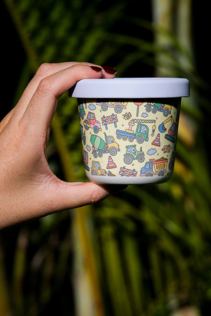 Hand holding BBCino reusable bamboo snack pot in Dig It design with colourful construction vehicles and lilac silicone lid, lightweight and durable.
