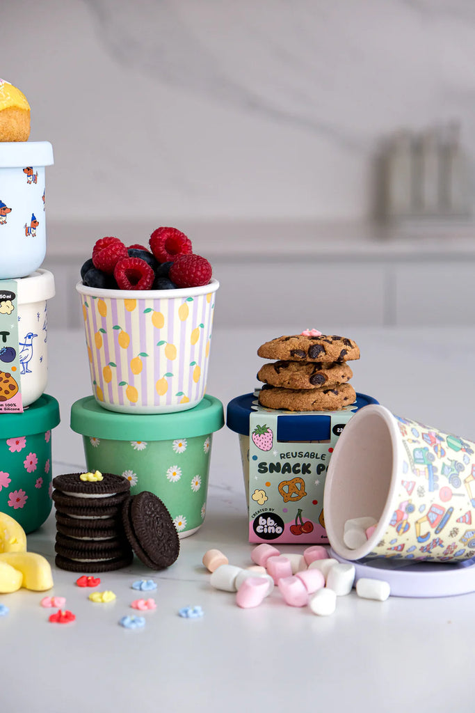 BBCino bamboo reusable snack pots in assorted designs including Dig It construction print, stacked on kitchen counter with snacks, eco-friendly containers.