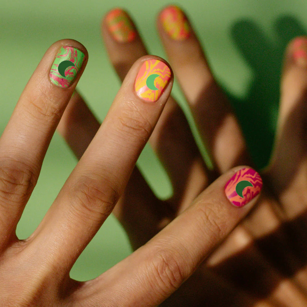 A close-up of fingers with bright, marbled nail art in pink, orange, and green, each nail decorated with a small green crescent moon sticker.