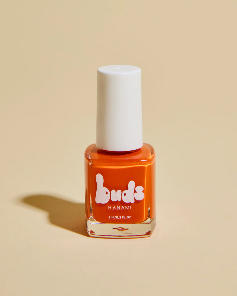 A bottle of Buds Hanami kids’ nail polish in a bright orange shade, standing on a beige background with a soft shadow