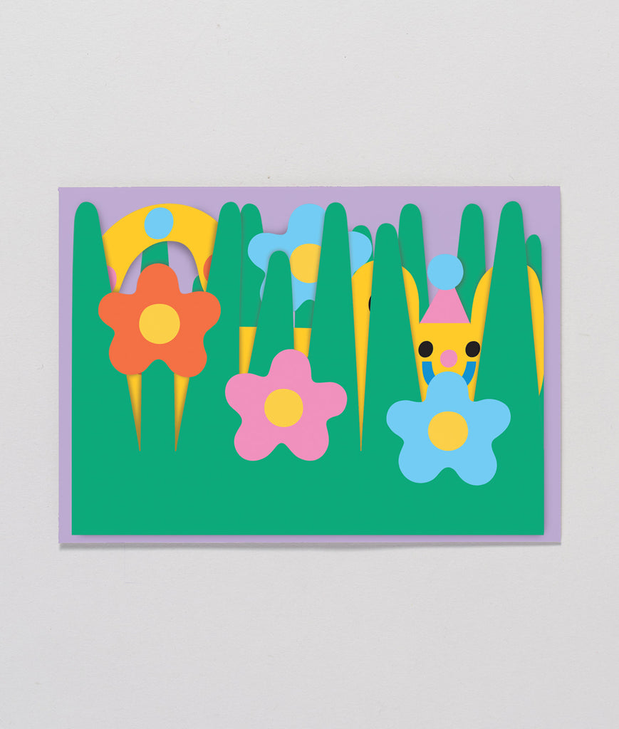 Flat lay of the cat fold-out birthday card fully closed displaying a colourful garden scene with flowers, grass, and a happy cat. Sitting atop the Lilac coloured envelope it comes with for gifting.