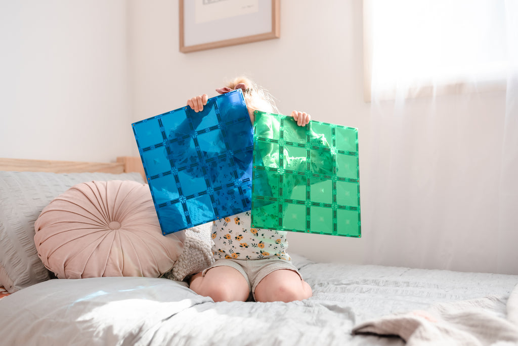 Child sitting on a bed holding the green Connetix base plate with the blue plate nearby, demonstrating scale and hands-on play.