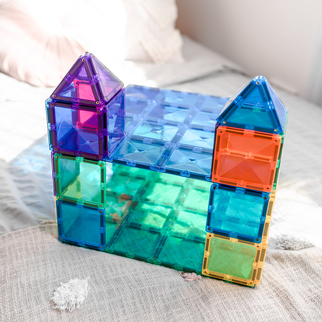 Magnetic tile castle built on top of the green and blue base plates, styled on a light-coloured bedspread with soft natural lighting.