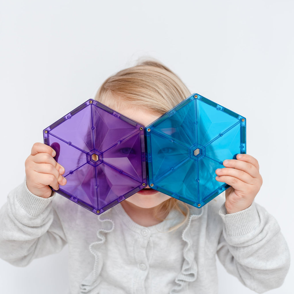 Child holding two large magnetic tiles over their eyes like glasses, smiling and playing with Connetix shapes.