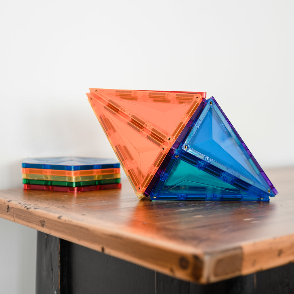 3D geometric pyramid built from rainbow magnetic tiles sitting on a wooden table, with a stack of additional tiles in the background.