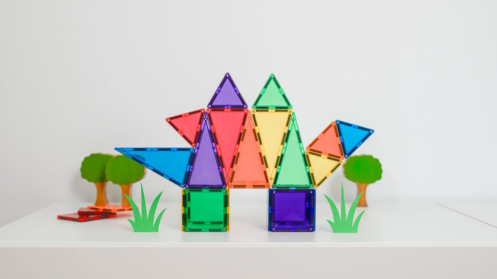 Creative dinosaur-style figure made from rainbow magnetic tiles, set on a white surface with small green tree props.