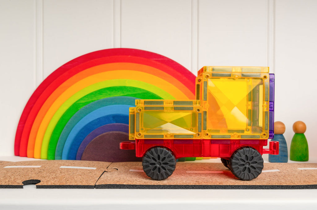 Yellow and red magnetic truck built using the Connetix Motion Pack, styled in front of a wooden rainbow stacker toy on a shelf.