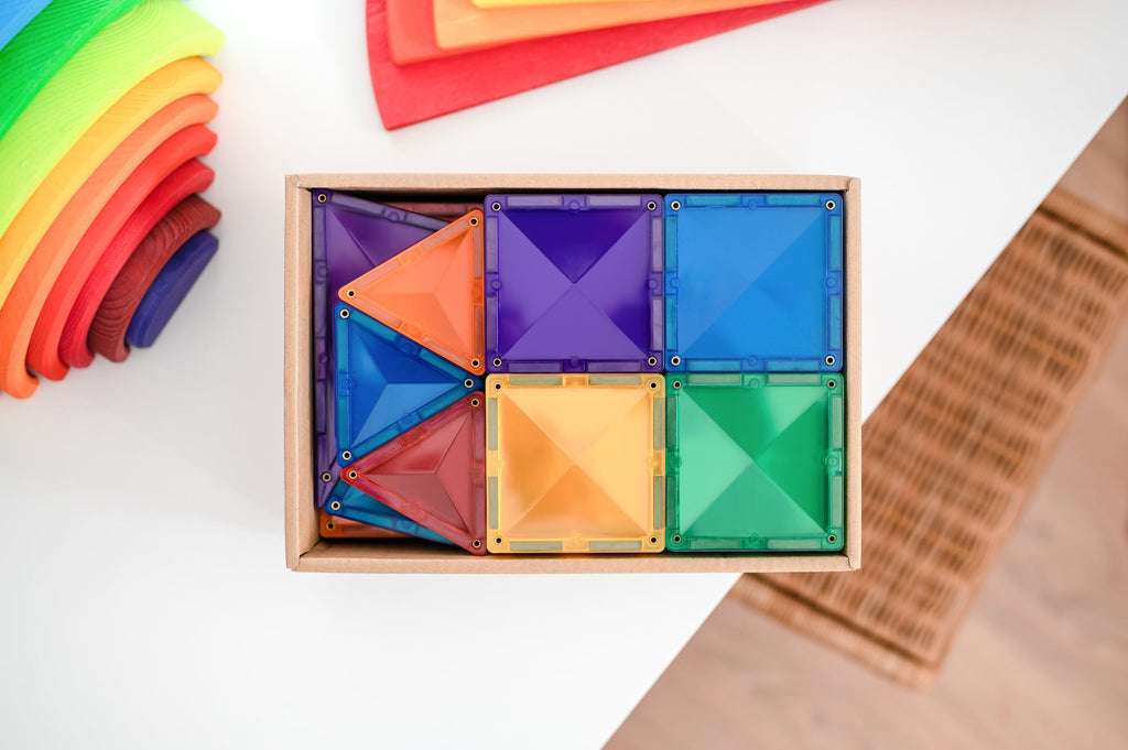 Top-down view of magnetic tiles neatly arranged by colour inside a wooden tray, next to a rainbow stacking toy.