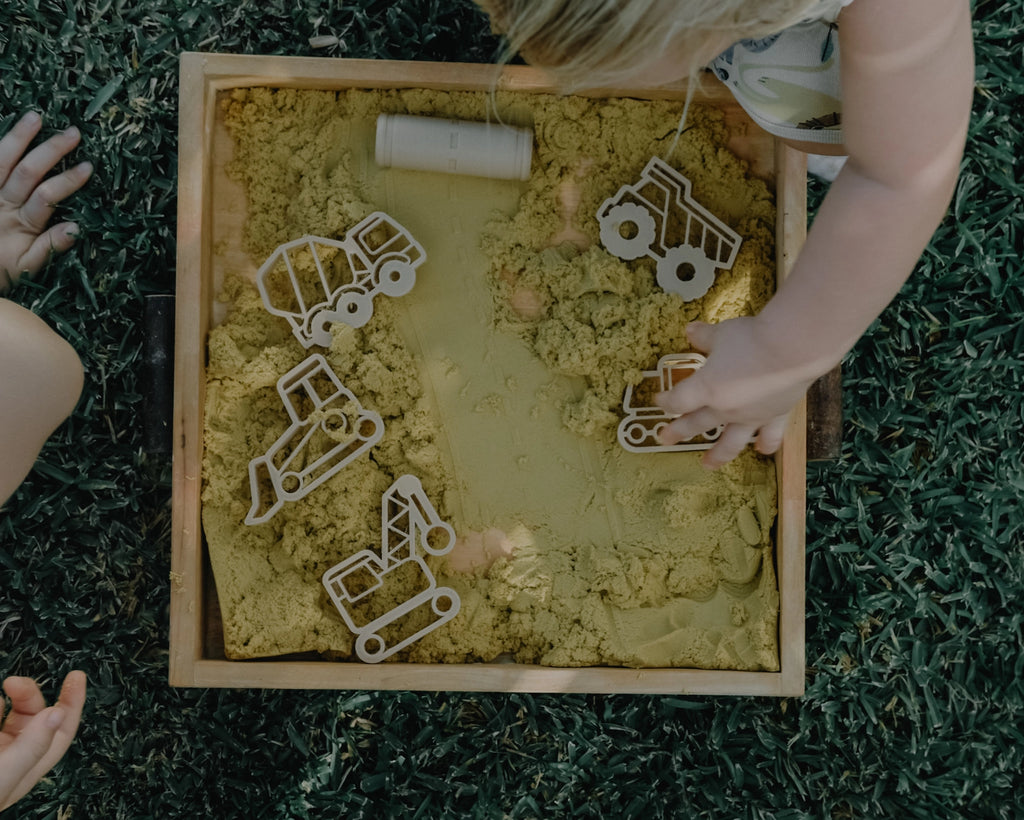 Children playing with sand and construction-themed toys in a sandbox outdoors.