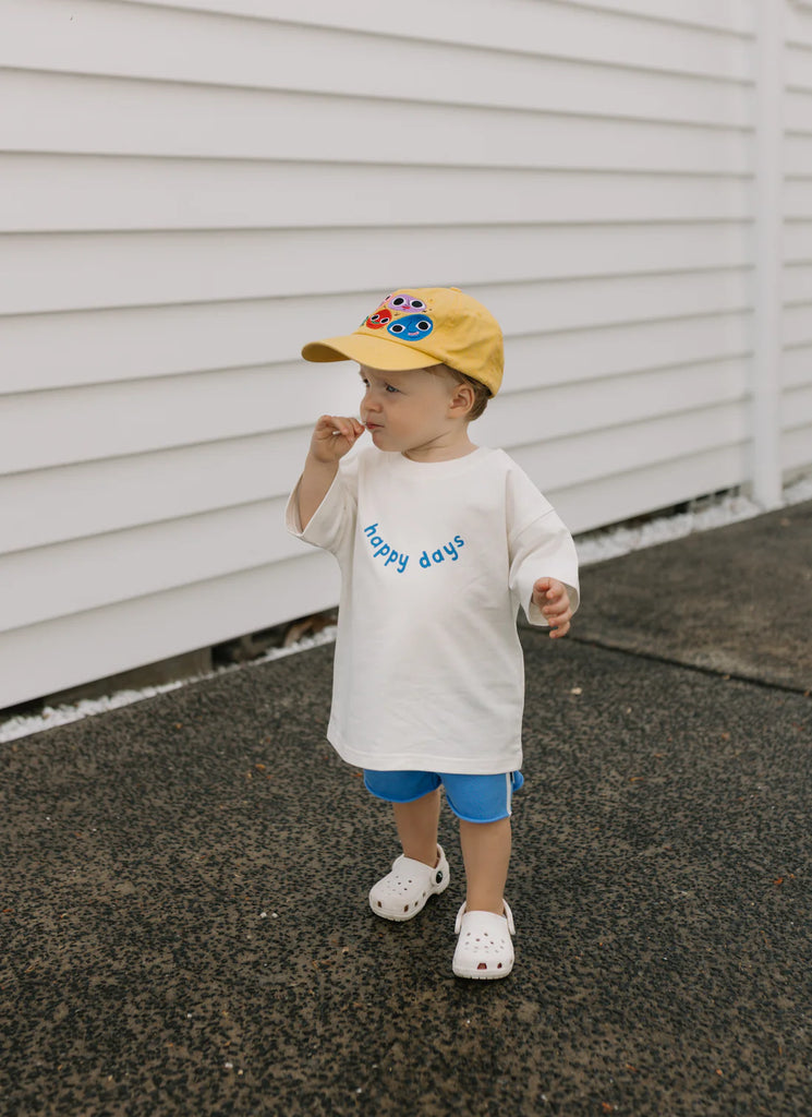 Child in Happy Days Tee, blue shorts, and a yellow cap standing confidently outside against a white weatherboard wall—ready for sunny adventures.