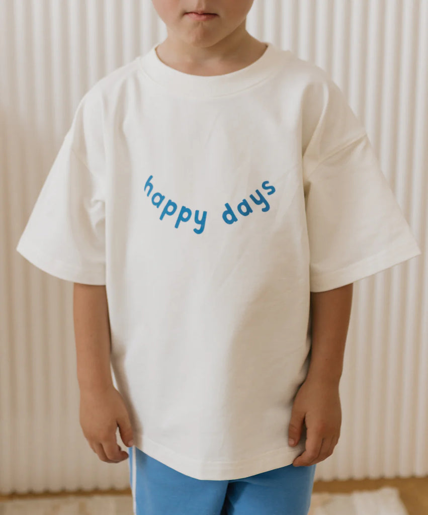 Child wearing the Happy Days Tee with blue lettering across the front, standing indoors and showcasing the oversized fit and soft cotton fabric.