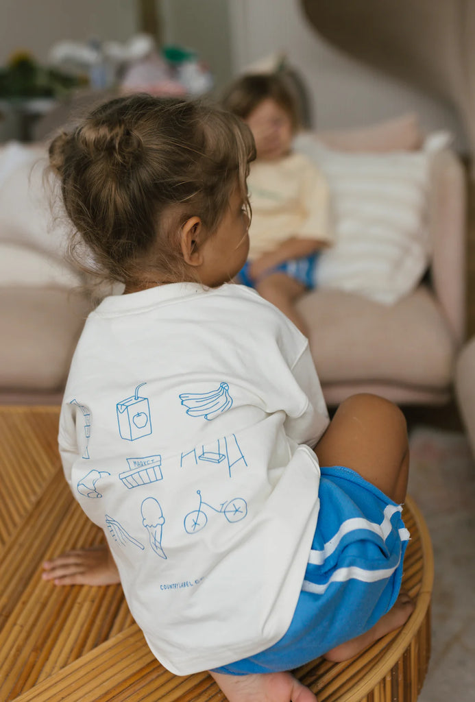 Child turned slightly to show the back of the tee, featuring whimsical hand-drawn illustrations in blue, highlighting the playful design element.