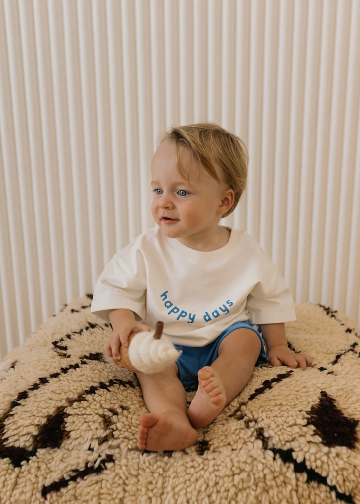 Young child sitting cross-legged on a round patterned rug, wearing the Happy Days Tee in a relaxed home setting with blue shorts.