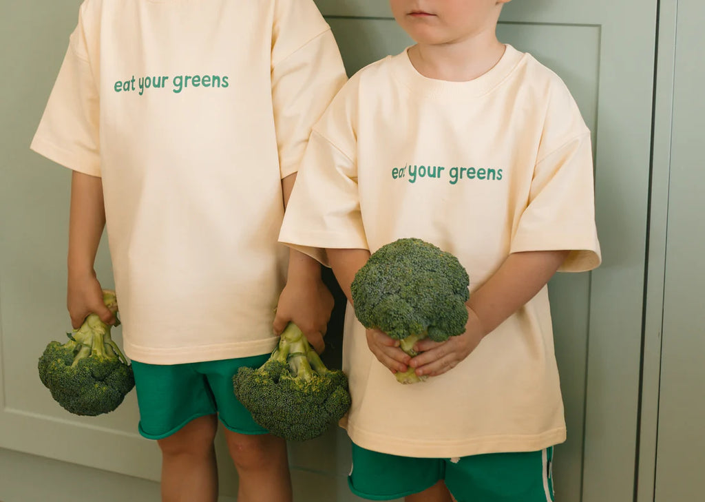 Two children wearing cream "Eat Your Greens" tees with green shorts, each holding a head of broccoli—highlighting the playful slogan and relaxed unisex fit.