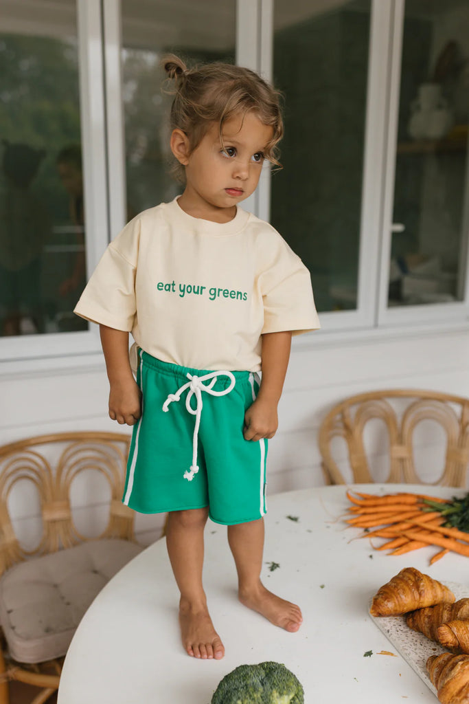 Toddler standing on a rattan chair wearing the Eat Your Greens tee and green racer shorts, showing the oversized fit and casual comfort of the outfit.