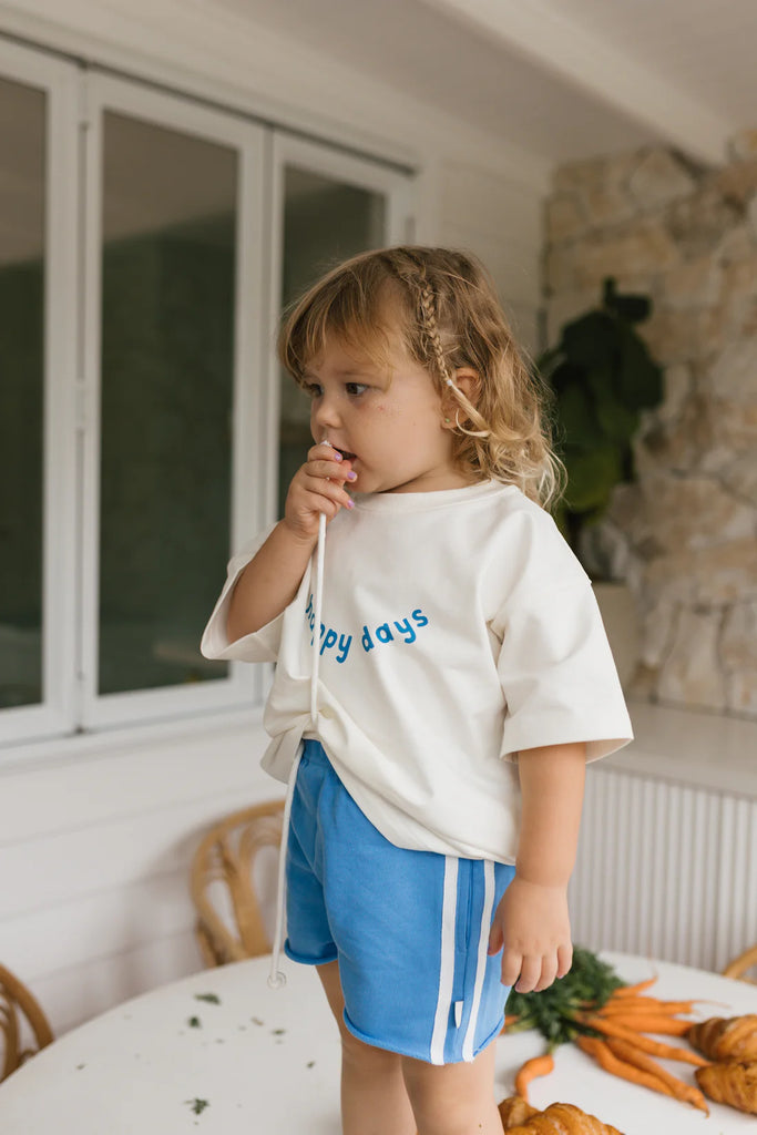 Child wearing bright blue racer shorts with white side stripes and a "happy days" tee, standing indoors in a relaxed setting, showcasing casual everyday wear.
