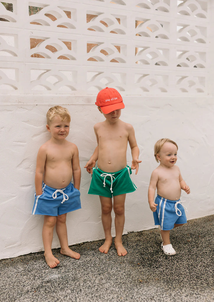 Three children standing outdoors wearing racer shorts in green and blue, displaying multiple colourways and a fun, coordinated summer look.