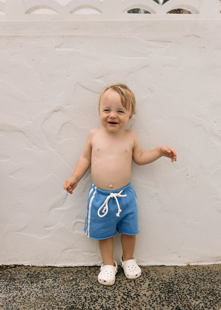 Child in blue cotton racer shorts smiling against a pale pink wall, barefoot and carefree, showing off the relaxed fit and drawstring waistband.