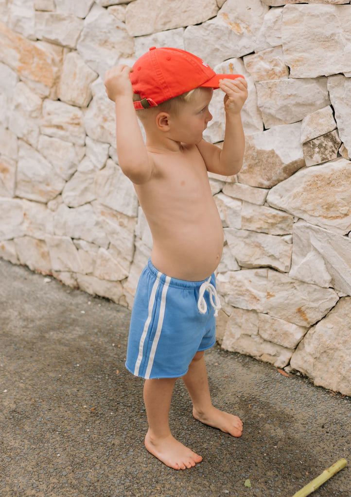 Toddler wearing blue racer shorts and a red cap, standing sideways against a textured stone wall, highlighting the sporty raw hem and elastic waistband.