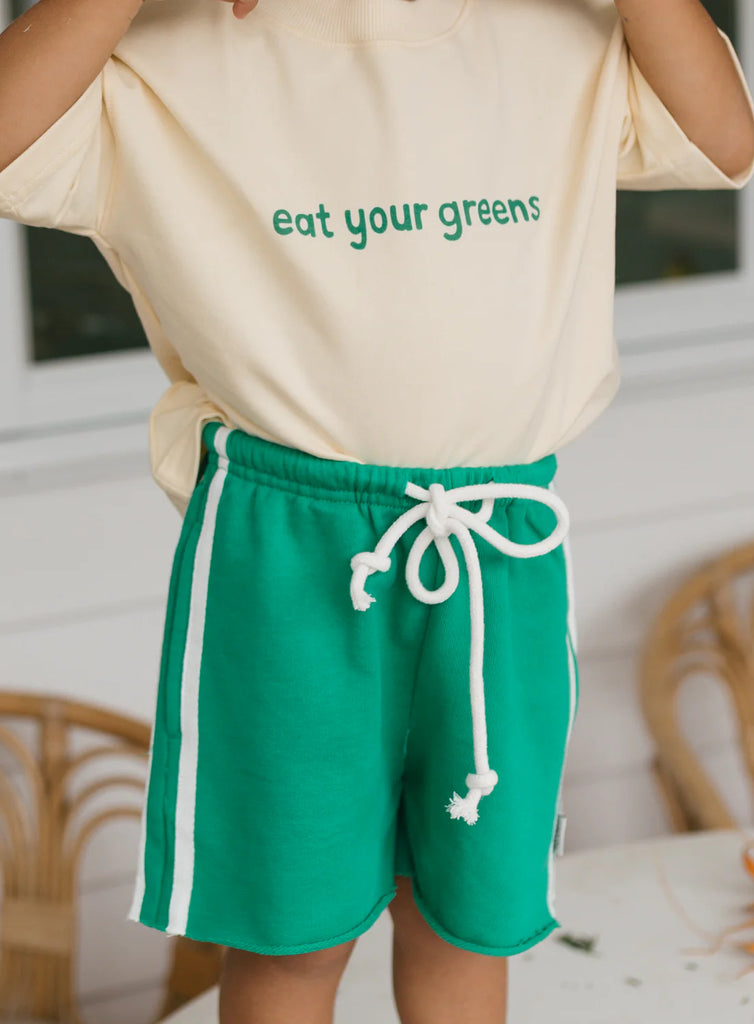 Child wearing vibrant green cotton racer shorts with a raw hem and white side stripe, paired with a cream "eat your greens" tee—perfect for casual summer play.