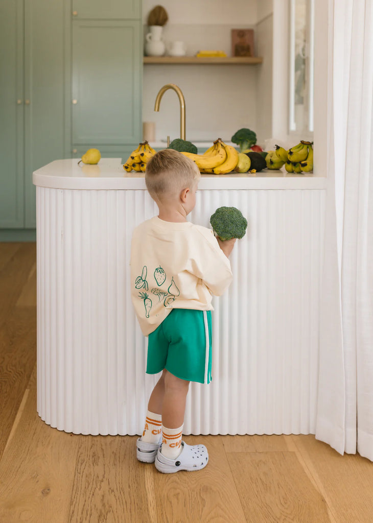 Young child in green racer shorts and a cream tee stands at a kitchen bench with lemons, showcasing the relaxed, playful fit and everyday wearability.