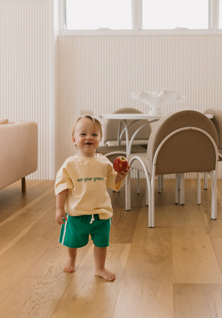 Toddler wearing green racer shorts playing indoors and eating an apple, highlighting the shorts’ flexibility, soft material and comfortable elastic waistband with drawstring.