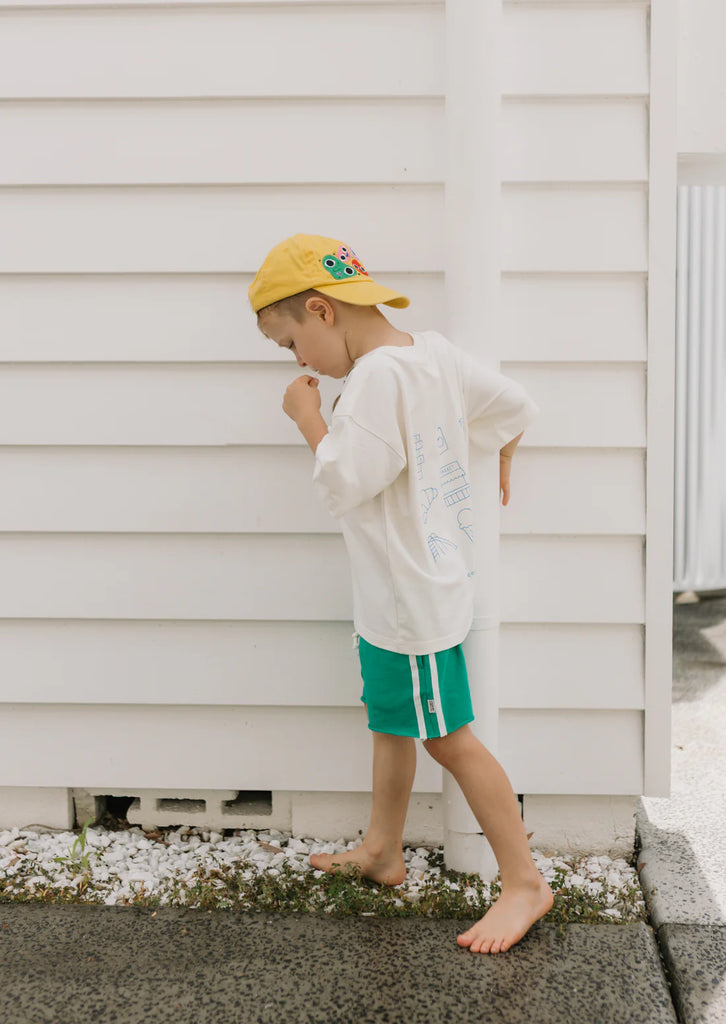 Child in green racer shorts and yellow cap standing barefoot against a white wall outdoors, demonstrating the shorts' durability and effortless cool look for summer days.