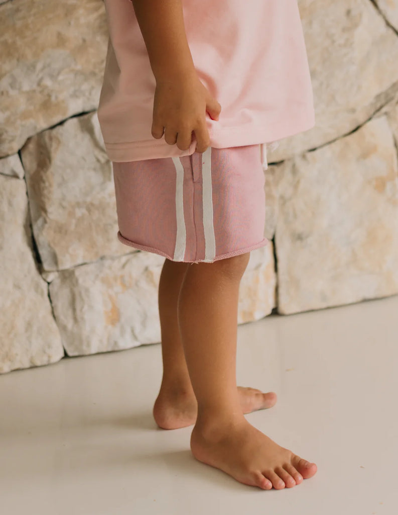 Close-up of child wearing soft pink racer shorts with raw hem and white side stripes, standing barefoot against a stone wall to highlight fabric and fit.