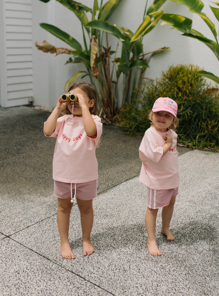 Two children outdoors wearing matching pink racer shorts and "play days" tees, smiling and posing side by side in sun hats, perfect for coordinated summer play.