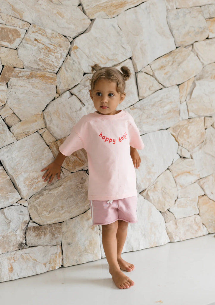 Child standing against a light stone wall wearing pink racer shorts and matching tee, capturing the playful, relaxed fit and sporty side stripe.