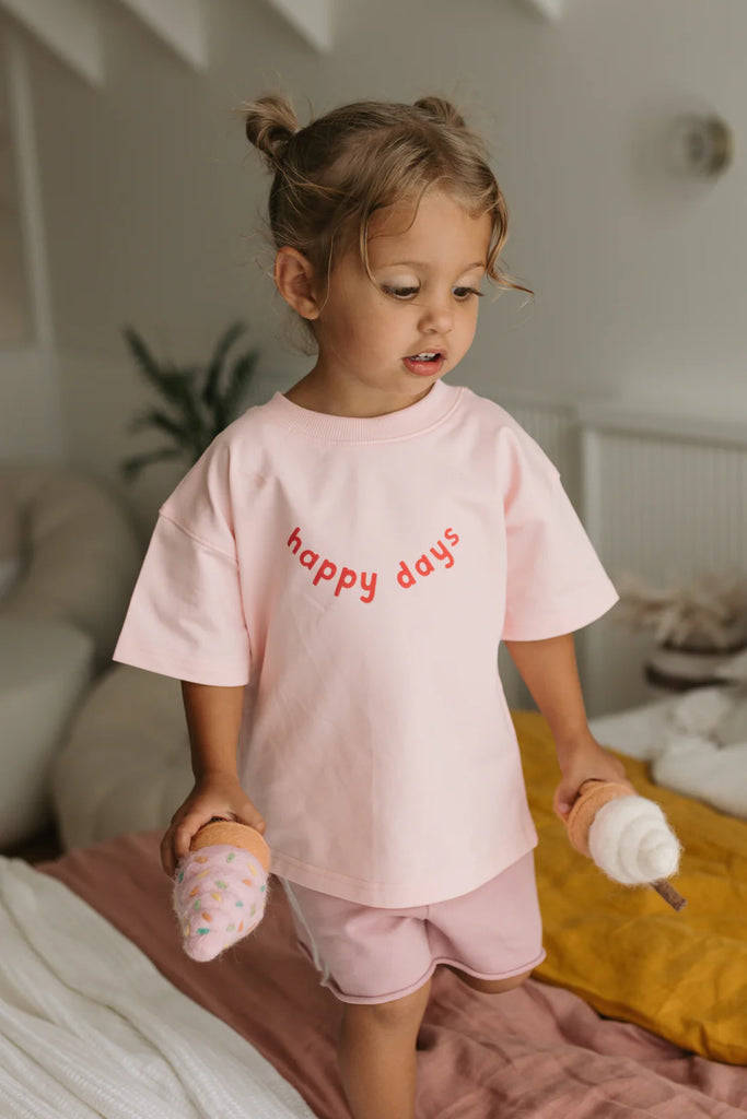 Toddler walking with a lollipop while wearing the Happy Days Tee in pink, showing the casual drape and bright red lettering on the front.