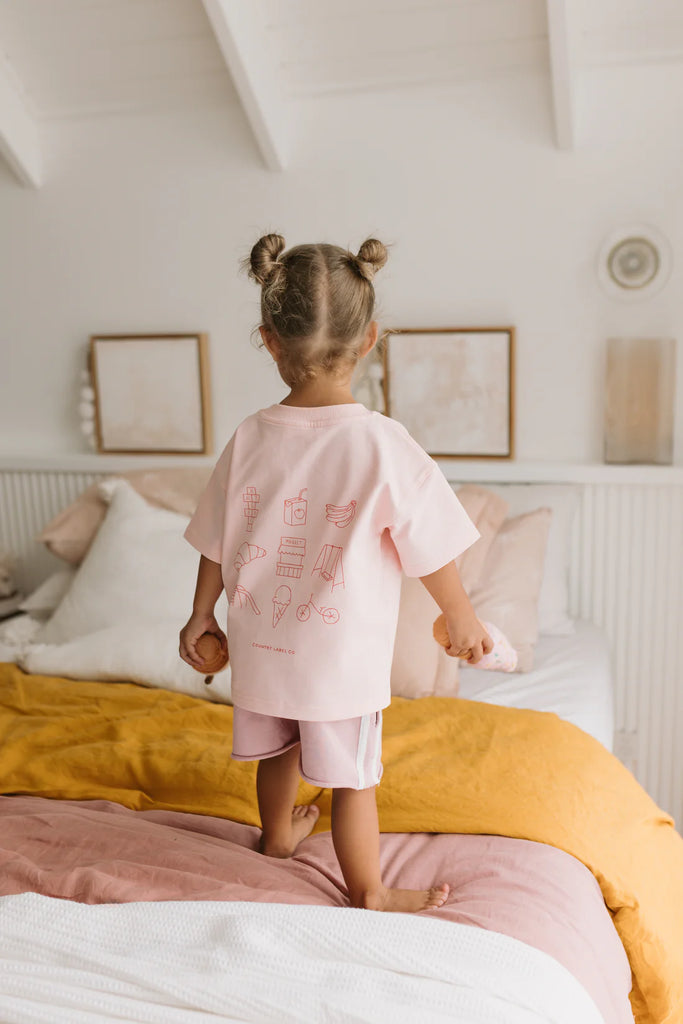 Child standing on a bed facing away, revealing the full back graphic of the pink Happy Days Tee with whimsical red line art.