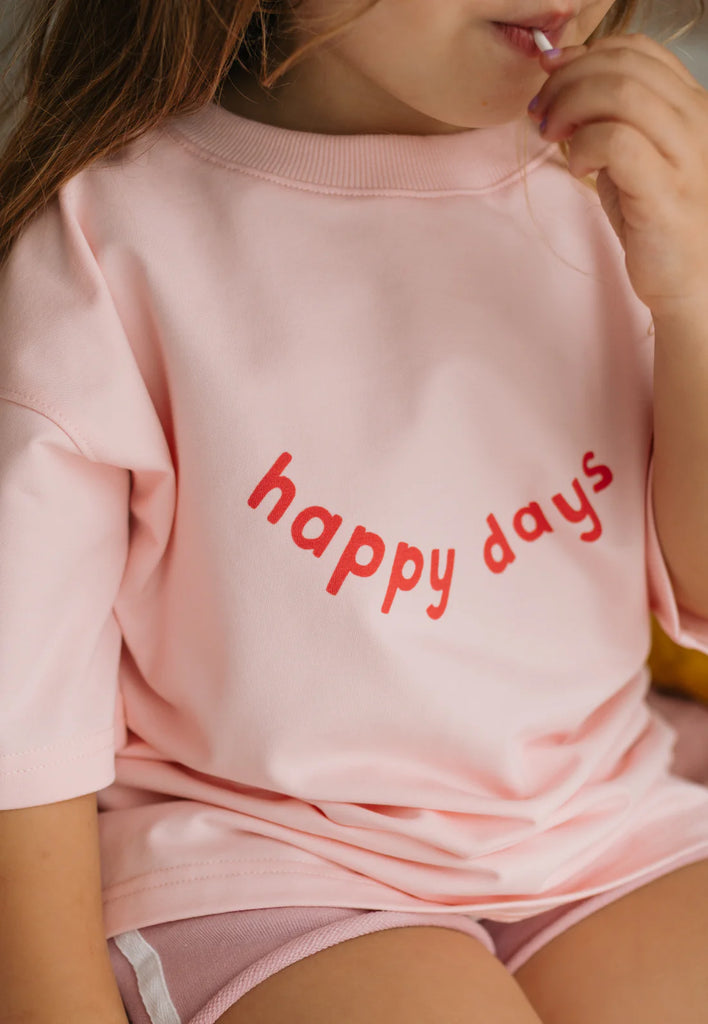 Close-up of a child wearing the soft pink Happy Days Tee with red “happy days” text on the chest, showing the smooth cotton fabric and playful slogan.