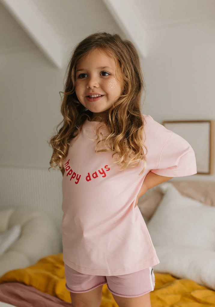 Child smiling while standing on a bed, wearing the pink Happy Days Tee and matching racer shorts, showcasing the relaxed fit and cheerful front design.